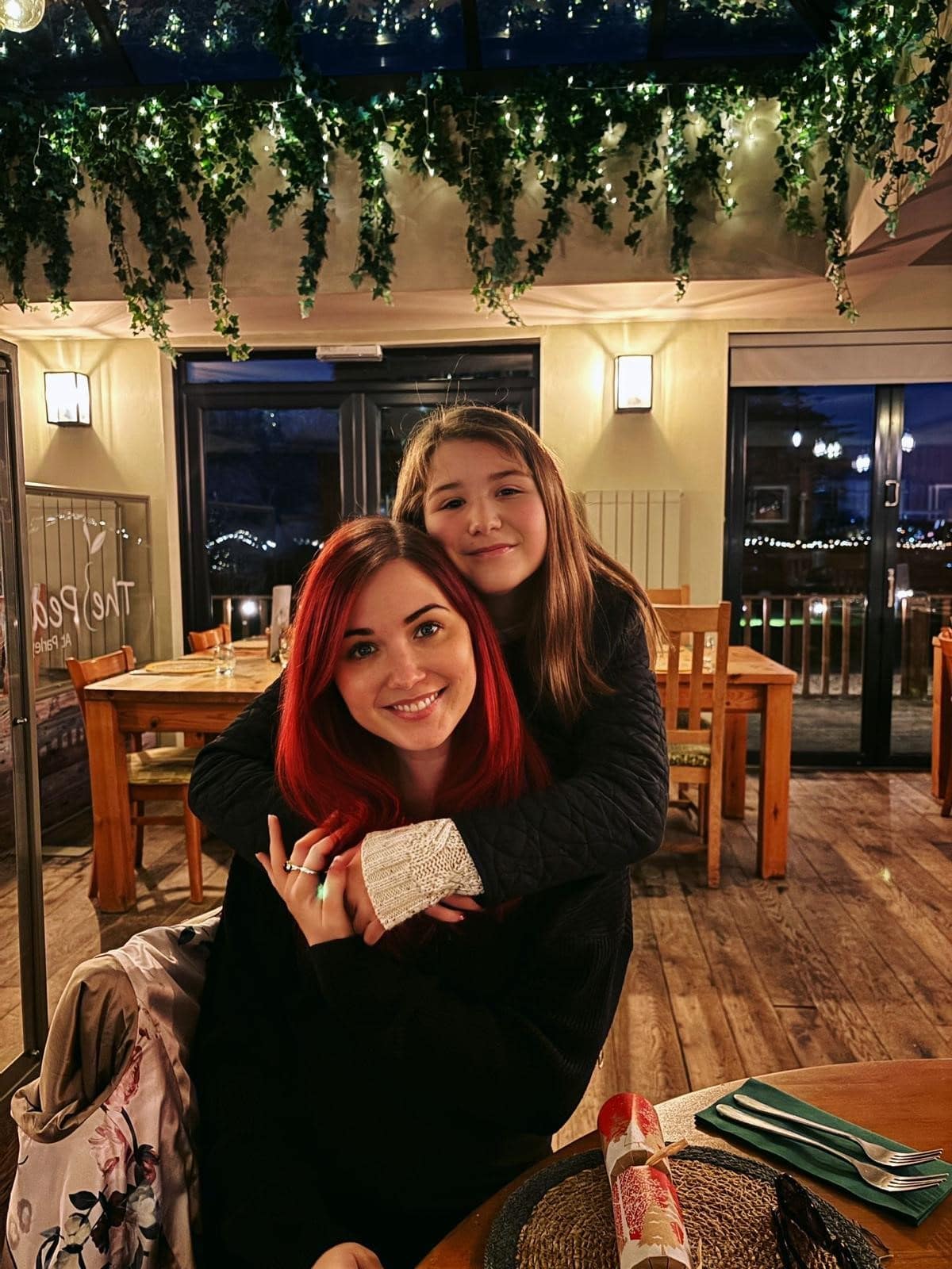 A woman, with long red hair, sits at a restaurant smiling at Christmas time. She is being hugged from behind by her teenage daughter