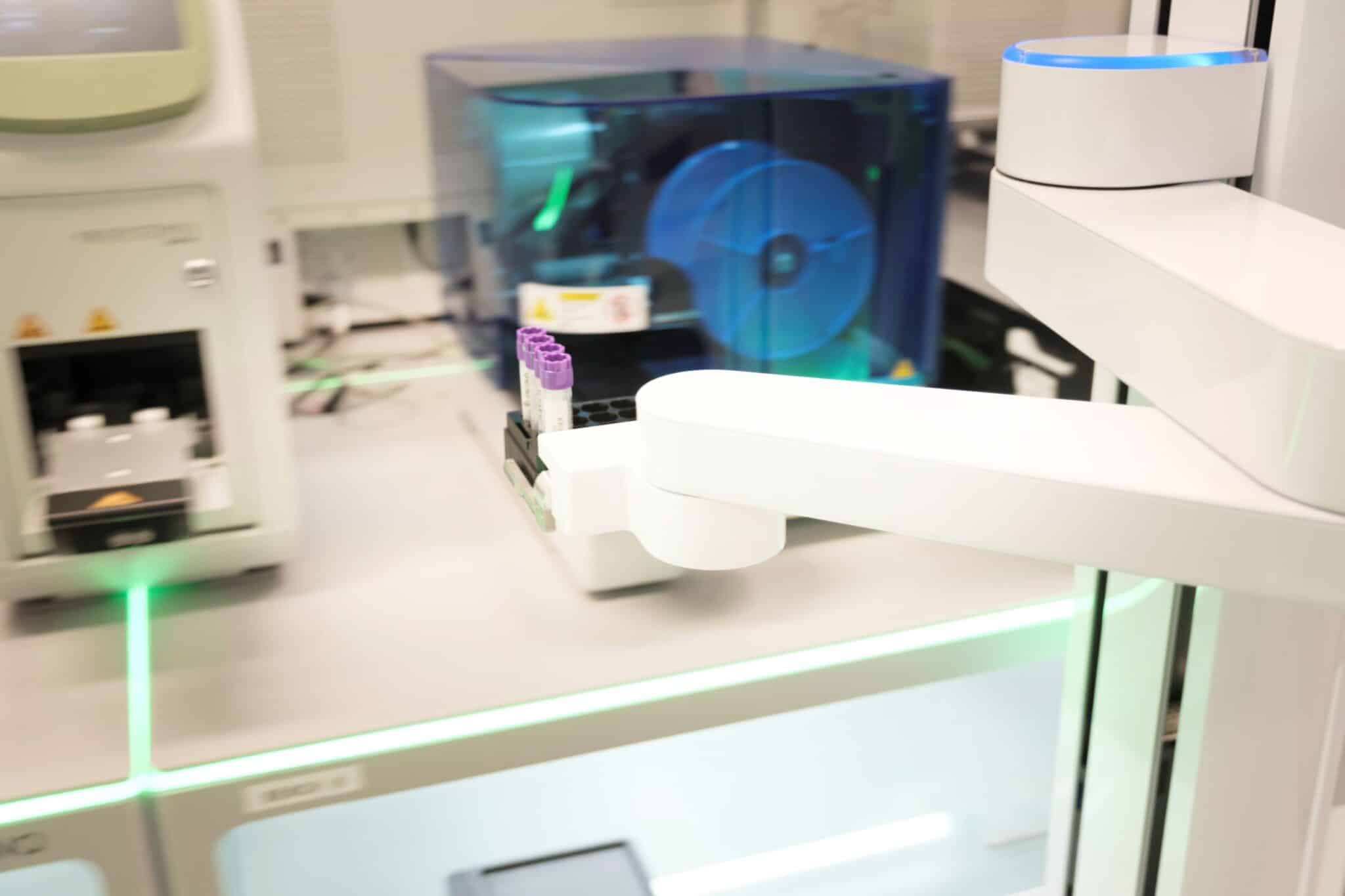 A close up shot of a piece of robotic lab equipment. The white moving robotic arm carries a tray with four purple topped test-tubes.