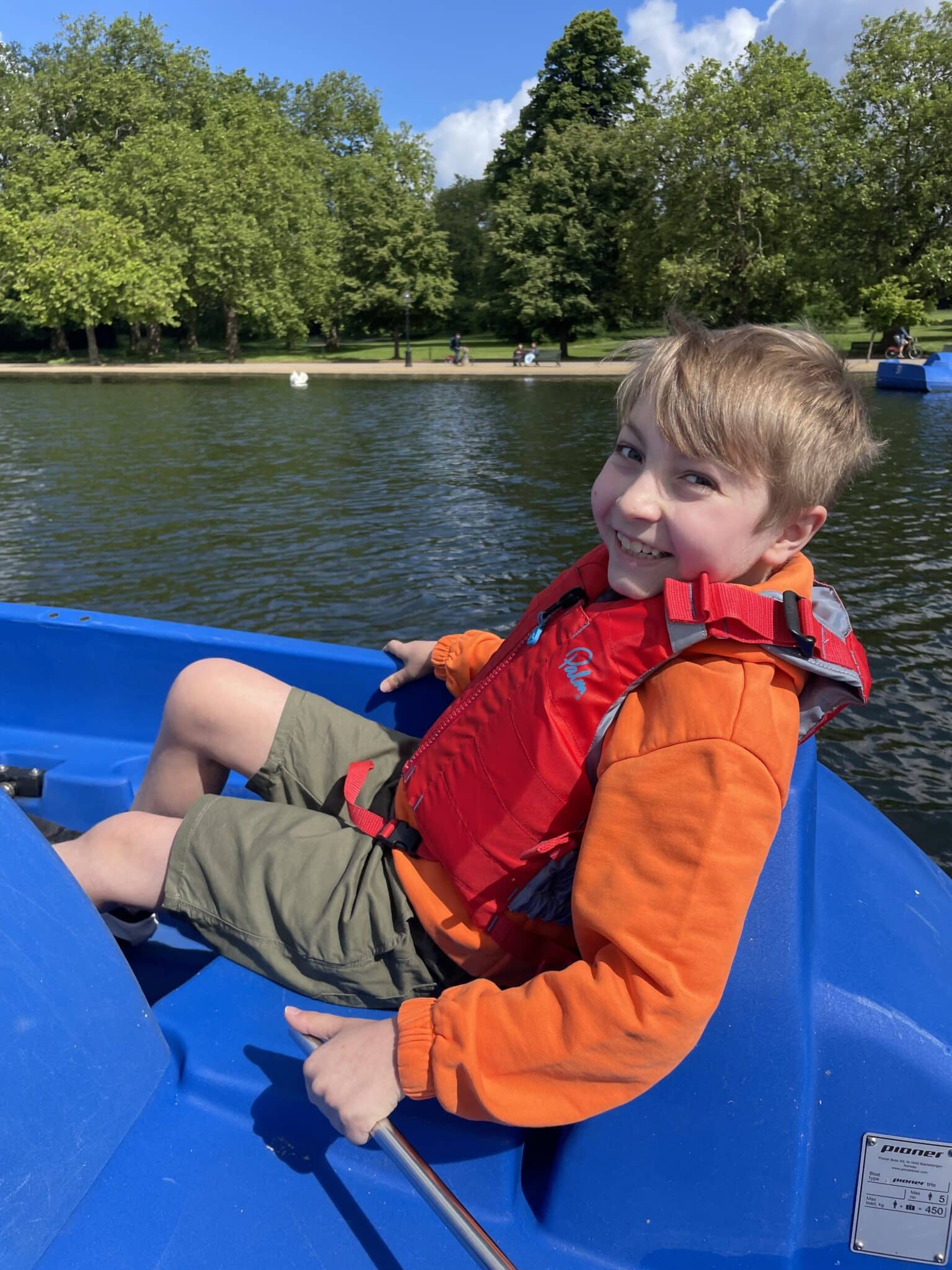 A boy in an orange jumper and a red life jacket as he smiles at the camera in a blue peddle boat