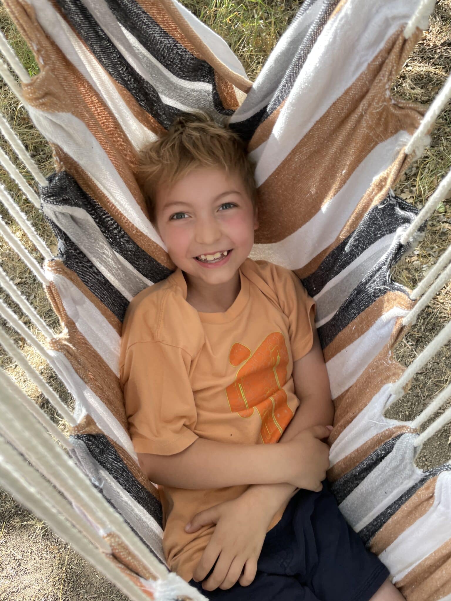 A young boy in an orange t-shirt lies in a hammock and smiles at the camera