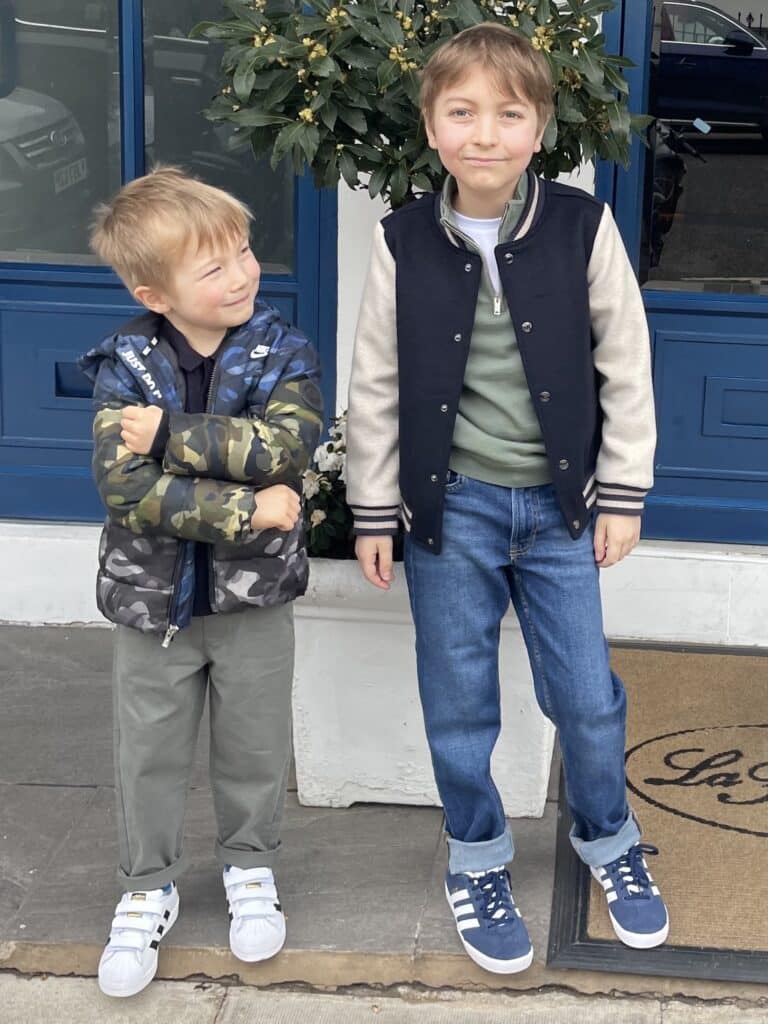 Two boys stand outside a restaurant in front of a plant. The smaller blonde haired boy wearing a camouflage jacket looks up at his older brother and smiles. His older brother stands to his right and smiles at the camera.
