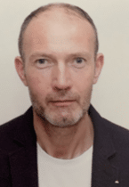 A man looks directly at the camera wearing a black blazer with a white t-shirt underneath